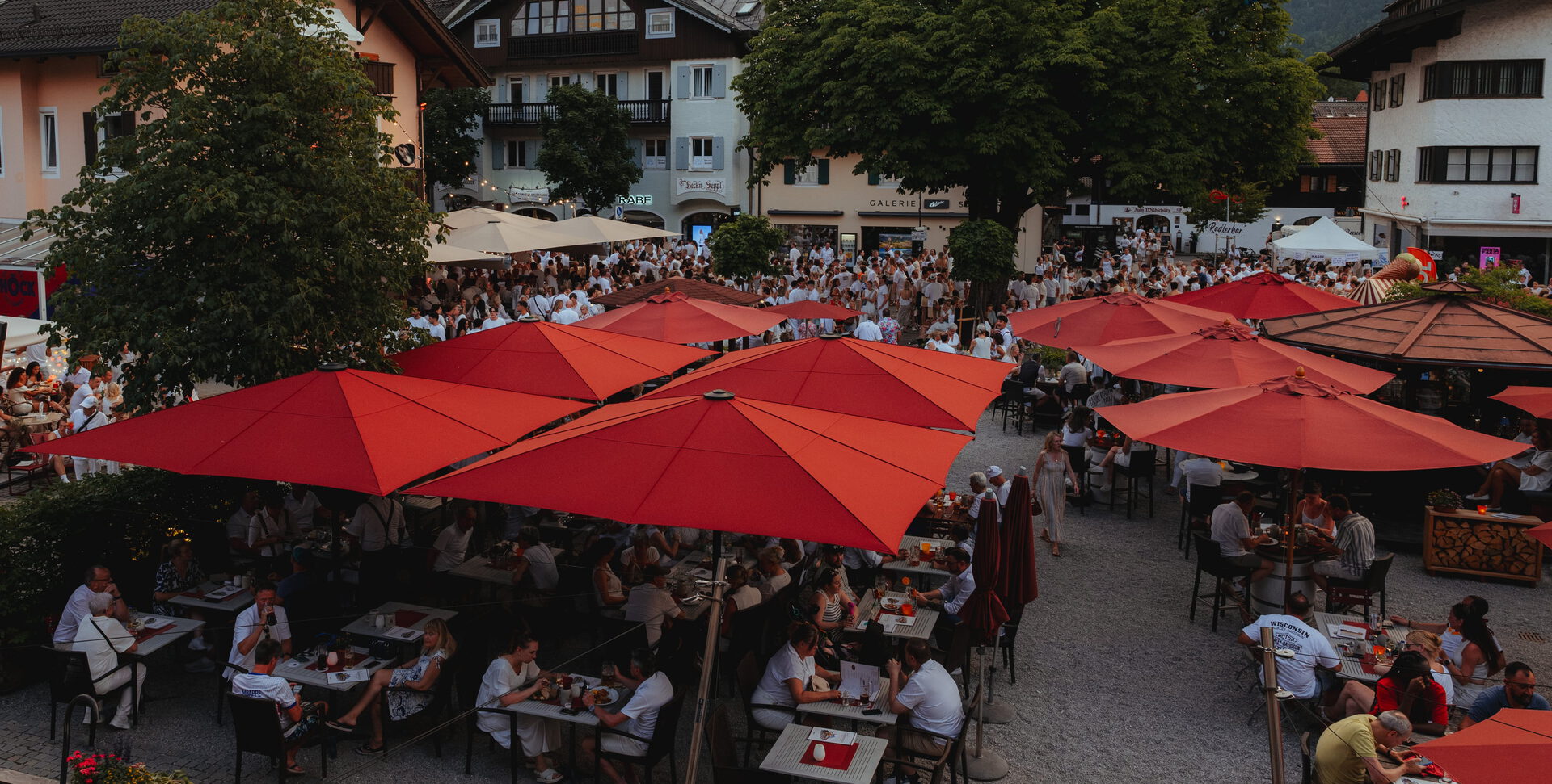 Blick von oben auf den vollen Biergarten unter roten Schirmen und einen Platz voller weiß gekleideter Festgäste
