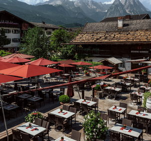 Biergarten mit roten Sonnenschirmen und gedeckten Tischen zwischen alten Häusern vor markanter Bergkulisse