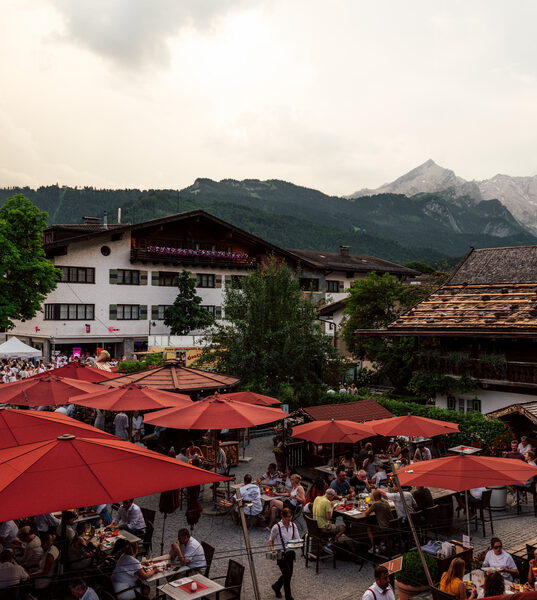 Kellner bedienen Gäste im gut besuchten Biergarten unter roten Schirmen, dahinter alte Häuser und Berge