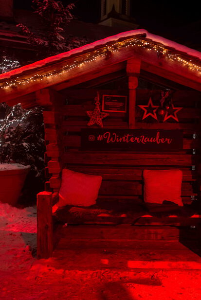 Rot beleuchtete Holzhütte mit Sitzbank und #Winterzauber-Schriftzug im Schnee