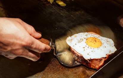 Hand wendet mit einem Pfannenwender eine gebratene Fleischscheibe mit Spiegelei auf der Grillplatte