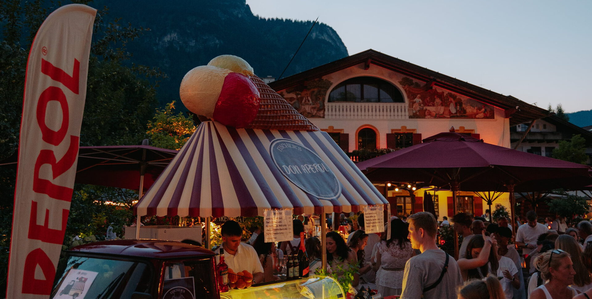 Beleuchteter Eisstand mit gestreiftem Dach und großer Eistüte am Abend, umringt von Gästen vor bemaltem Haus