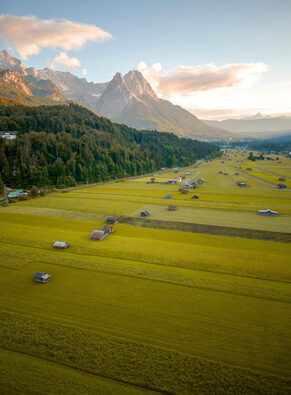 Luftaufnahme weiter Wiesen mit vielen Heustadeln in einem Tal, dahinter Wald und schroffe Berggipfel