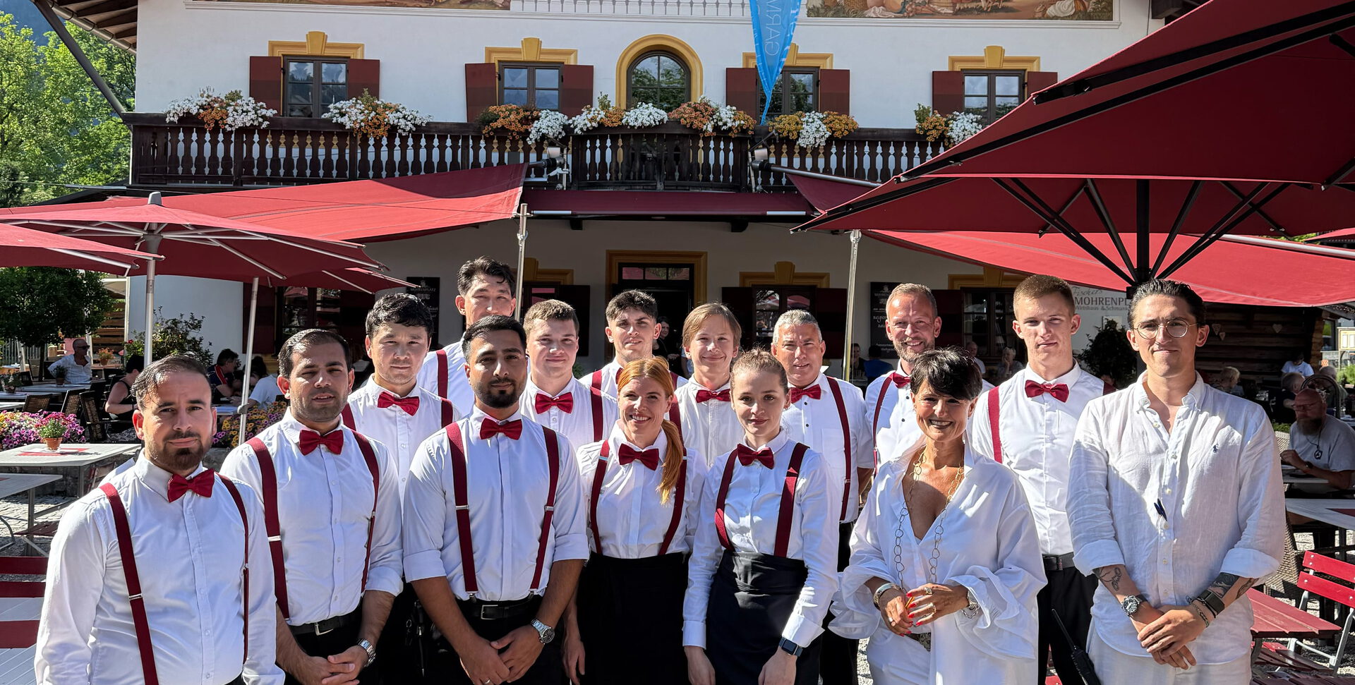 Gruppe von Servicekräften mit roten Fliegen und Hosenträgern vor dem bemalten Wirtshaus unter roten Schirmen