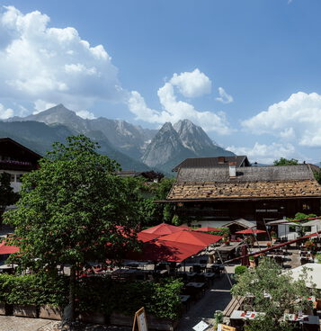 Blick von oben auf den Biergarten mit roten Schirmen neben bemaltem Wirtshaus und Zwiebelturm vor Bergpanorama