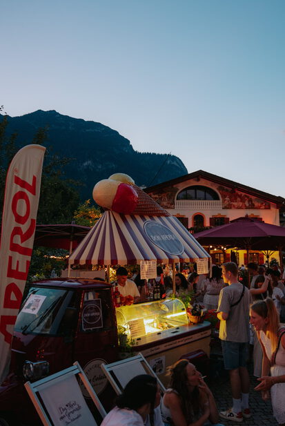 Beleuchteter Eisstand mit gestreiftem Dach und großer Eistüte am Abend, umringt von Gästen vor bemaltem Haus