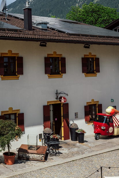 Weißes Wirtshaus mit Fensterläden, Kirchturm und Bergen, davor Terrasse und ein rotes dreirädriges Eismobil