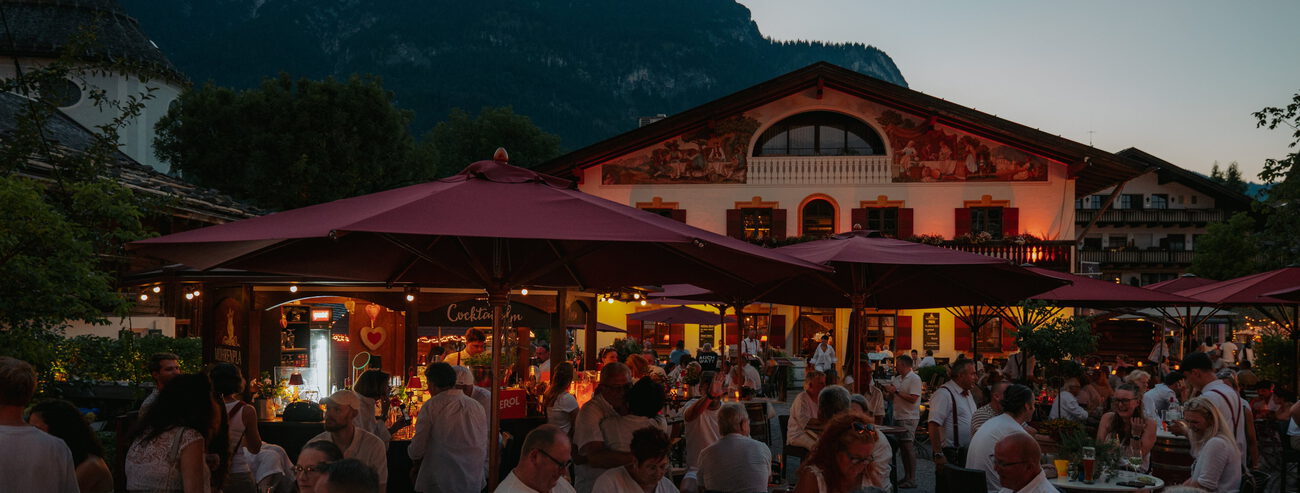 Voll besetzter Biergarten in der Abenddämmerung unter dunkelroten Schirmen vor bemaltem Wirtshaus und Bergsilhouette