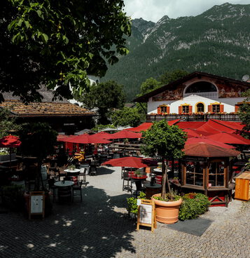Schattiger Biergarten mit roten Schirmen, Holzbar und Liegestühlen vor bemaltem Wirtshaus und bewaldetem Berg