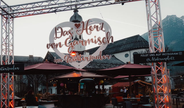 Torbogen mit Leuchtherz „Das Herz von Garmisch im Winterzauber“ vor Stehtischen, Schirmen, Kirchturm und Bergen