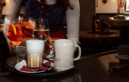 Bedienung in Dirndl trägt ein Tablett mit Spritz, Weißwein, Latte macchiato und Steinkrug „Mohrenplatz Garmisch“