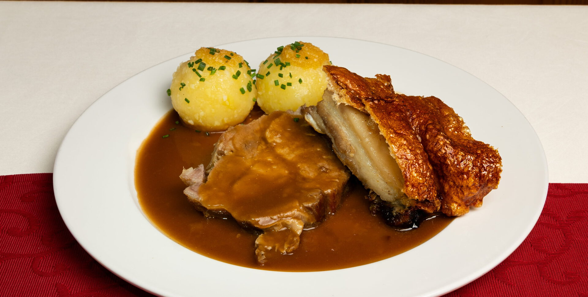 Braten mit knuspriger Kruste, zwei Kartoffelknödeln mit Schnittlauch und dunkler Soße auf weißem Teller