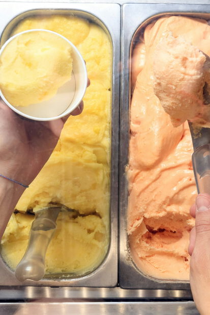 Hände portionieren Speiseeis aus einer Eistheke mit weißen, gelben, orangen und dunklen Sorten in einen Becher