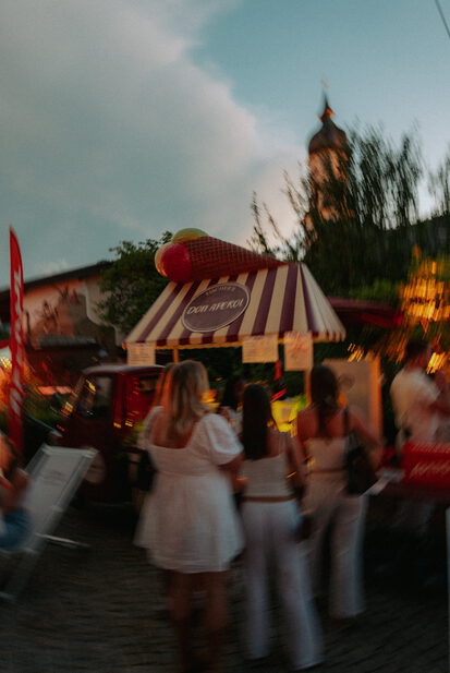 Verwischte Abendszene mit weiß gekleideten Gästen vor einem gestreiften Stand mit Schild „Don Aperol“ und großer Eistüte