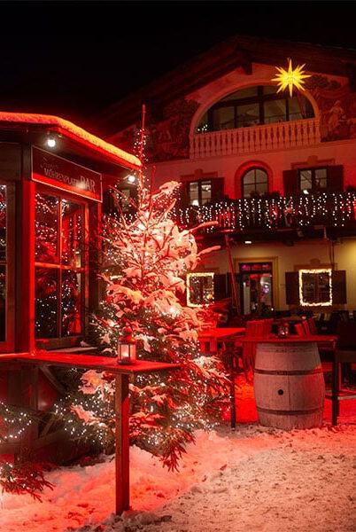 Verschneite, beleuchtete Tannen vor einer Holzhütte mit Aufschrift „Bar“ und dem weihnachtlich geschmückten Wirtshaus bei Nacht
