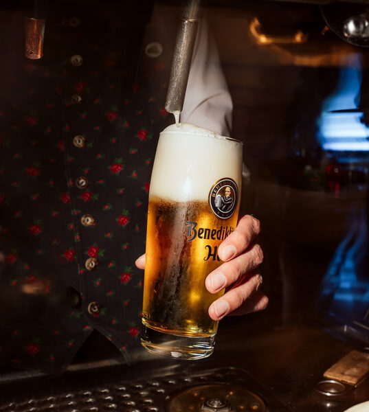 Bier wird in ein Glas mit der Aufschrift „Benediktiner“ gezapft, dahinter eine geblümte Trachtenweste