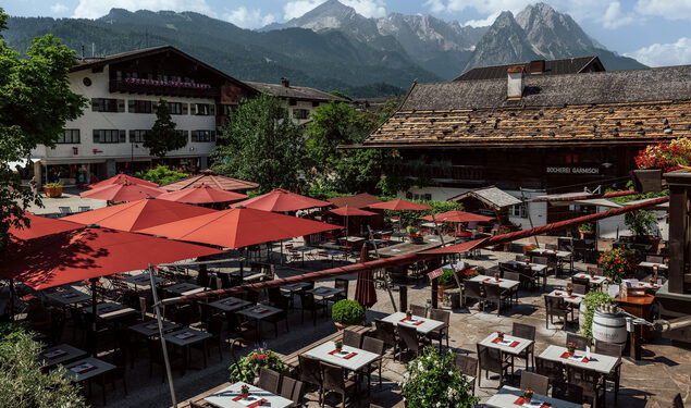 Biergarten mit roten Sonnenschirmen und gedeckten Tischen zwischen alten Häusern vor markanter Bergkulisse