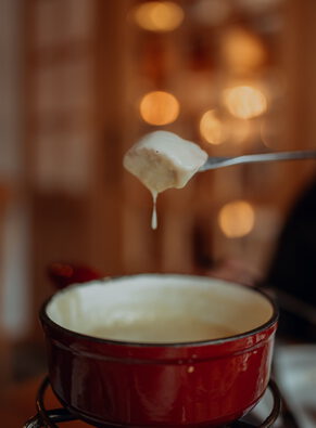 Brotwürfel an einer Fonduegabel tropft über einem roten Käsefonduetopf, dahinter warme Lichtpunkte