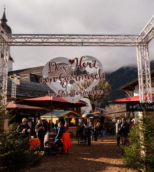 Torbogen mit Schild „Das Herz von Garmisch im Winterzauber“, darunter Gäste an Stehtischen vor Kirchturm und Wirtshaus