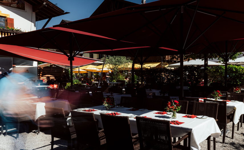 Festlich gedeckte Tafeln mit weißen Tischdecken, roten Servietten und Blumen unter roten Schirmen im Biergarten