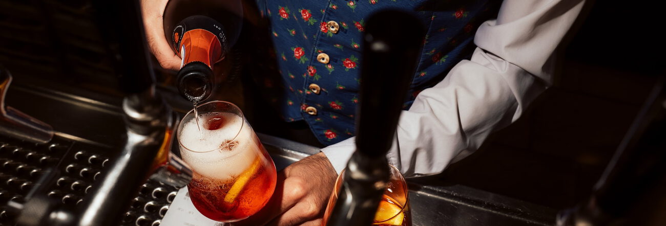 Kellner in geblümter Trachtenweste gießt Sekt in ein Glas mit orangerotem Spritz an der Schanktheke