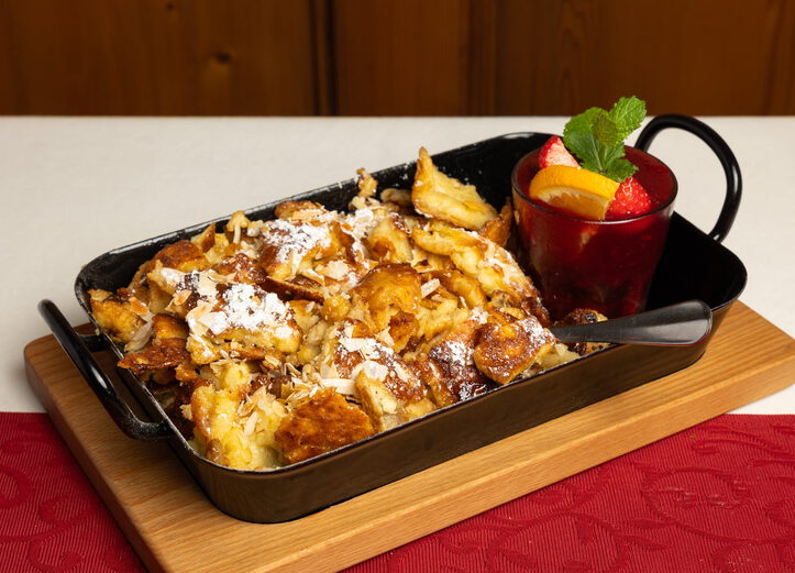 Kaiserschmarrn mit Mandelblättchen und Puderzucker in einer Pfanne, daneben ein Glas Fruchtkompott mit Minze