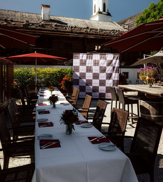 Lange weiß gedeckte Tafel mit roten Servietten und Blumen im Freien, dahinter Fotowand, altes Holzhaus und Kirchturm
