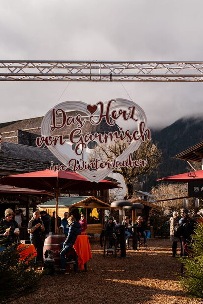 Torbogen mit Schild „Das Herz von Garmisch im Winterzauber“, darunter Gäste an Stehtischen vor Kirchturm und Wirtshaus