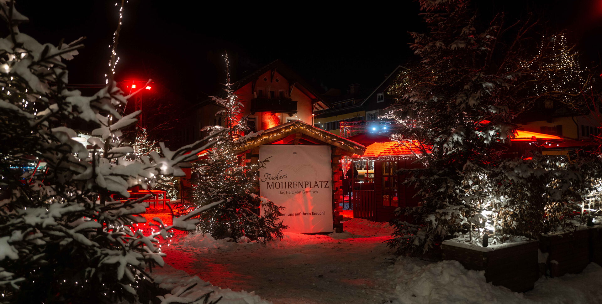 Verschneiter Eingang bei Nacht mit beleuchteten Tannen und Schild „Fischers Mohrenplatz – Das Herz von Garmisch“