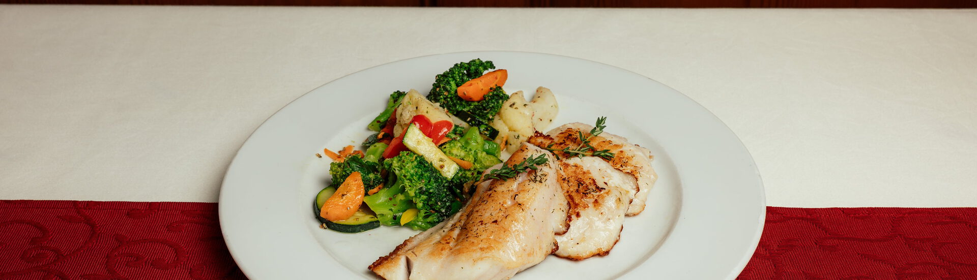 Gebratenes Fischfilet mit Thymian und buntem Gemüse auf weißem Teller vor einer Holzwand